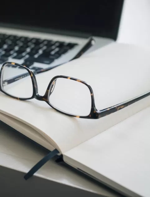 Glasses laying on a notebook beside a pencil and a laptop
